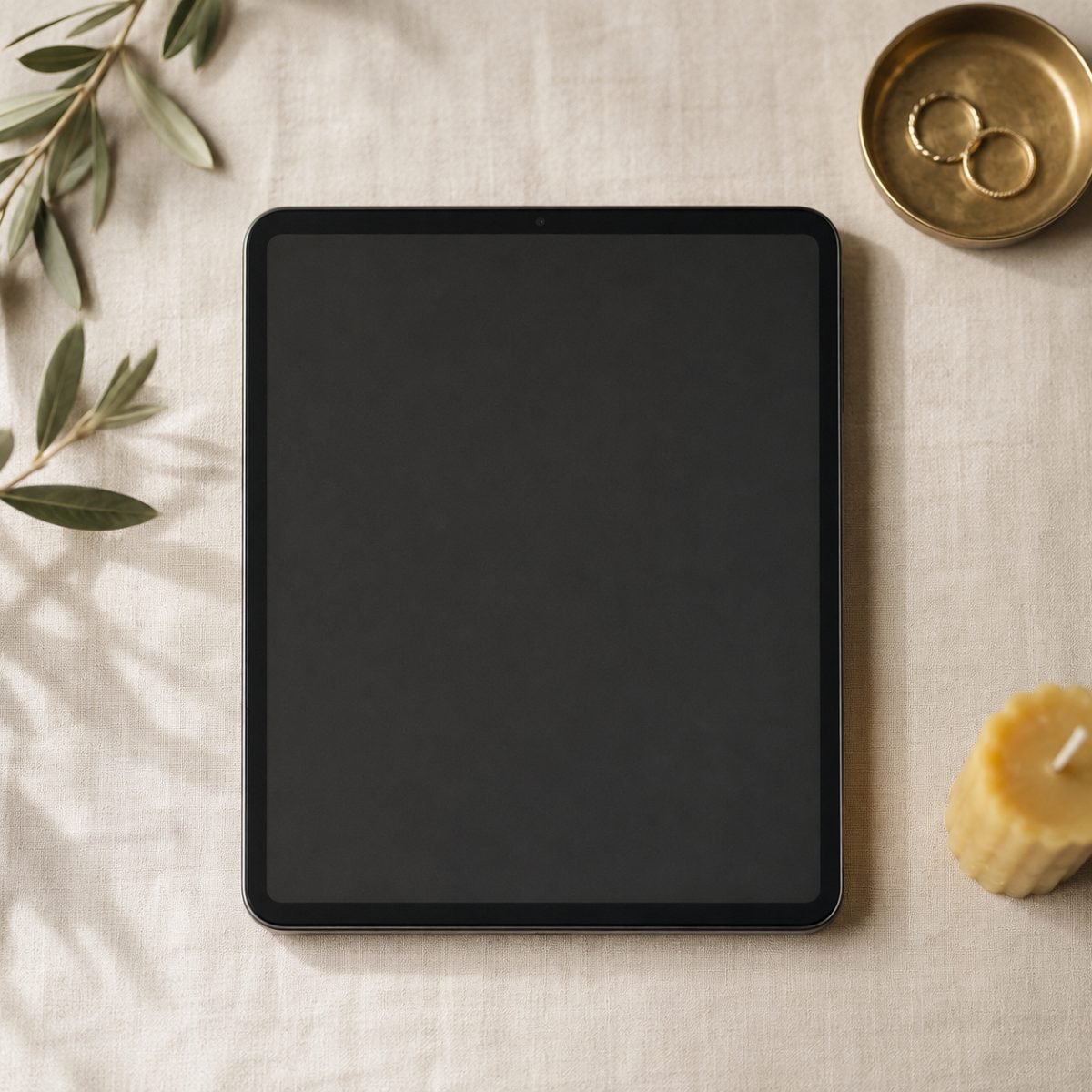 A tablet showing a wedding website home page, on cream linen with olive branches, a brass ring dish and a beeswax candle