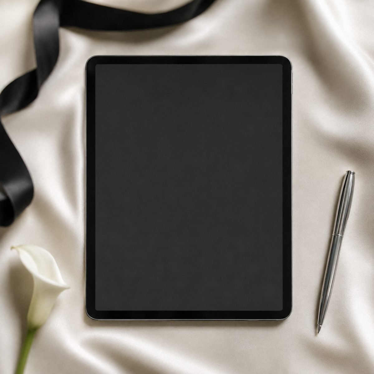 A tablet showing a wedding RSVP page, on ivory silk with a black ribbon, a silver pen and a white calla lily